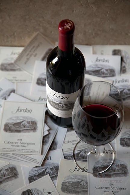 Jordan Cabernet Sauvignon bottle with a poured glass on a table with multiple vintages of wine labels.