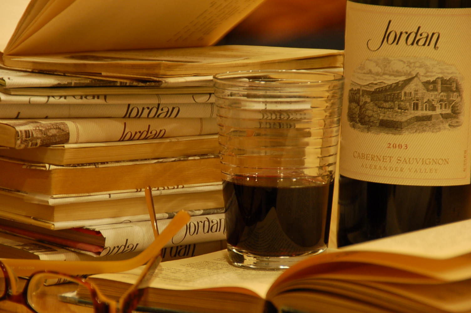 "Wine Literacy" a photo of a bottle of 2003 cabernet next to a poured glass surrounded by stacks of books covered with Jordan Winery labels by Jordan Catapano - Jordan Winery Celebrating Family with Jordan photo contest grand prize winner