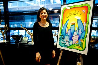 Andrea Arroyo posing next to her entry piece "New York Celebrates Jordan" which was awarded second runner up at 4 on 4 New York. She has long black hair and a long sleeve black shirt. Her art features a vibrant blue curvy cityscape. "Jordan" is written in cursive with a bird against a yellow sunset background. A dancing teal woman and green leaves make up the bottom corners. There's a squiggly bright green border.