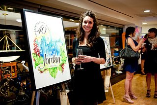Stephanie Mead posing next to her art piece "Celebrate!" which won grand prize at the 4 on 4 New York. She has long brown curly hair and is wearing a black dress and cardigan. Her art features the word "Jordan" supported by grape leaves and purple grapes. Above "Jordan" are vibrant blue glass buildings and "40" on the top left.
