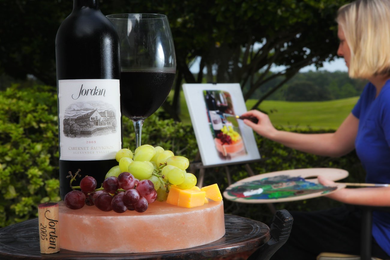 "Drink and paint!" a photo of a bottle of Jordan Winery cabernet on a wheel of cheese next to some table grapes and a poured glass of red wine with a woman painting a portrait of it in the background - What Would You Pair with Jordan Wine photo contest grand prize winner