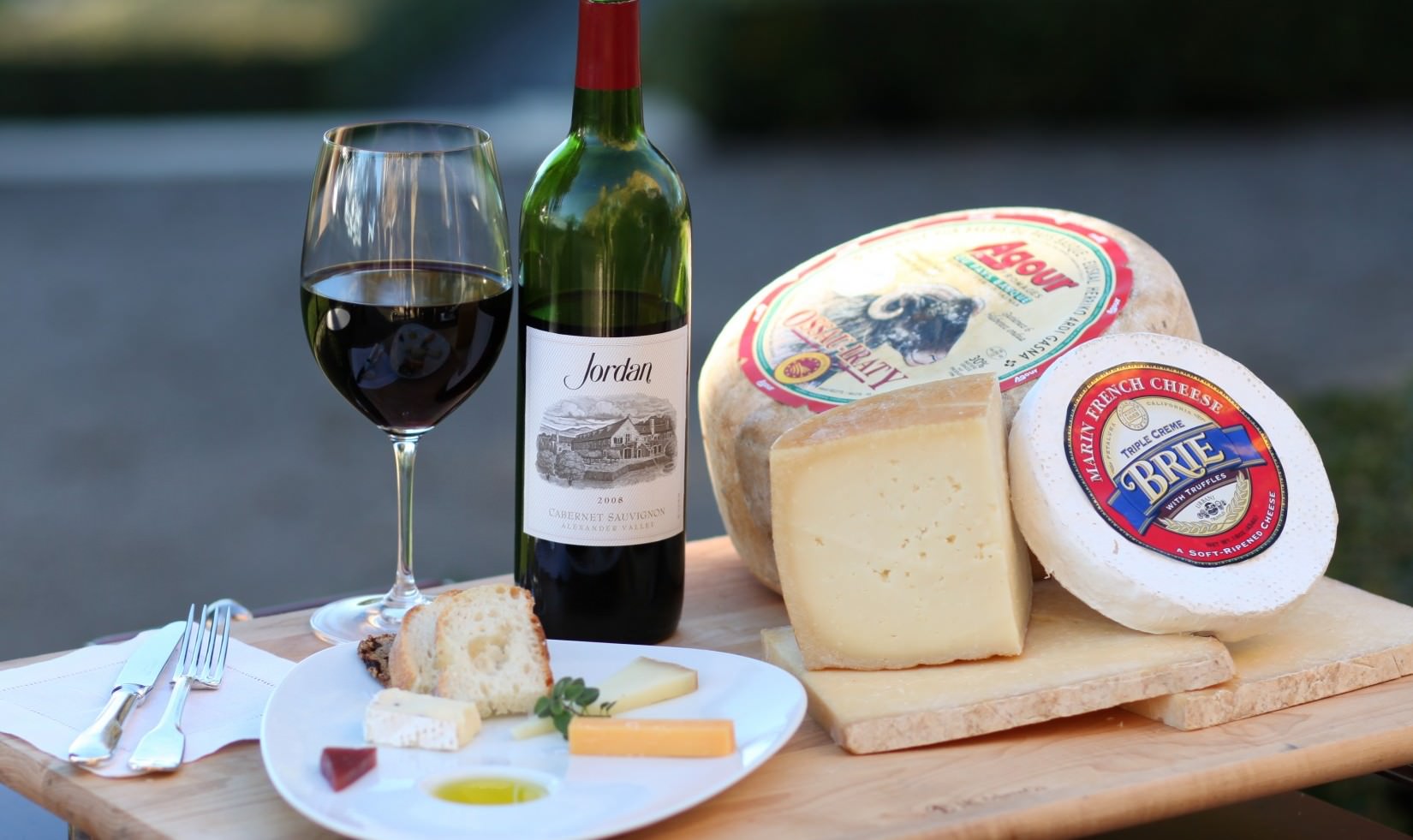 wheels of Agour Ossau-Iraty and Marin French Cheese Triple Cream Brie next to a cheese plate, a bottle of Jordan Winery cabernet and a poured glass of cabernet