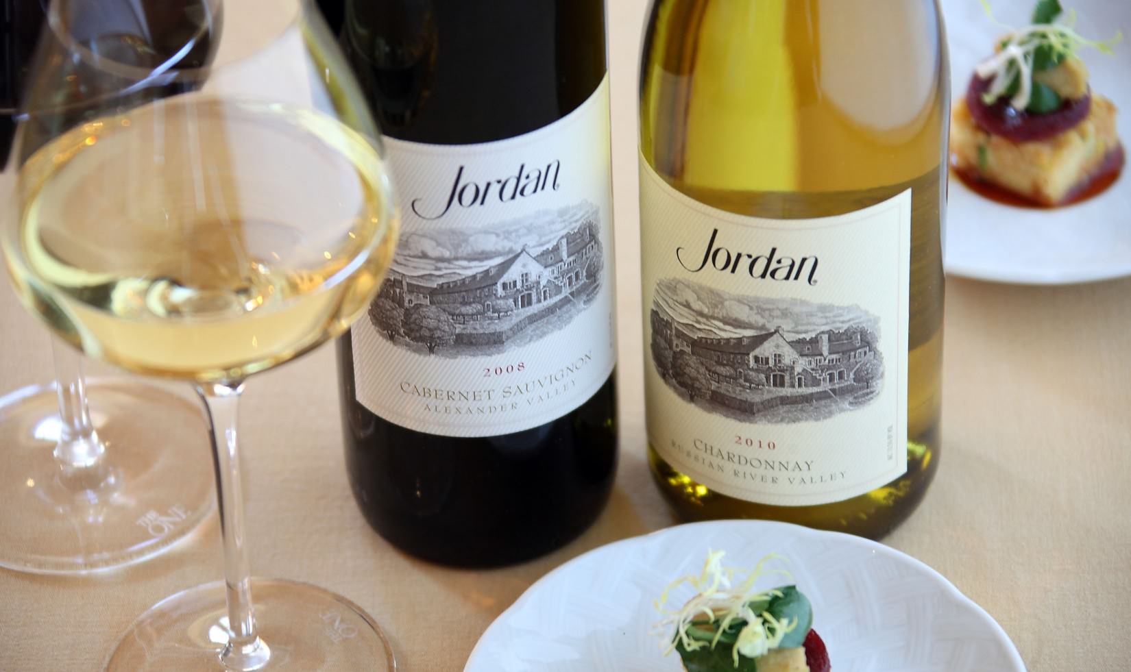 a bottle of 2008 Jordan Winery Cabernet and a bottle of 2010 Jordan Winery Chardonnay next to a poured glass of Chardonnay and small appetizers on small plates