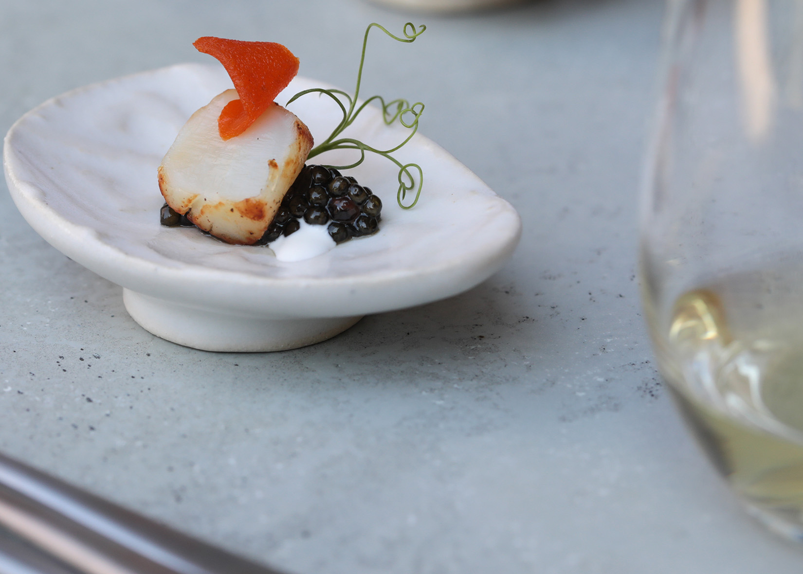 Bay scallop with pickled persimmon skin and caviar on dish next to wine glass