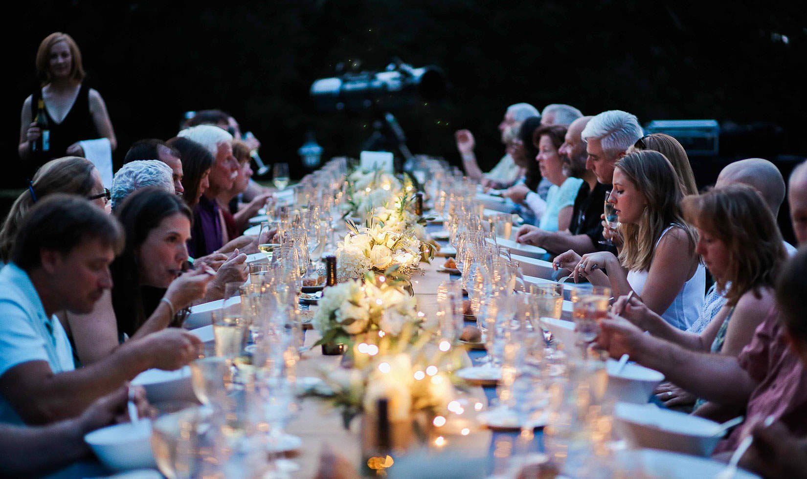 Guests seated at a large alfresco dining table at the Starlight Supper: Perseids Stargazing event. Candles and small white bouquets line the table.
