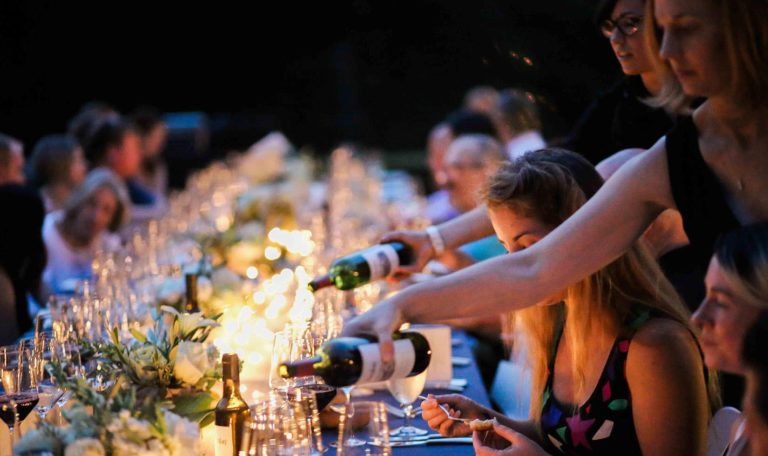 Guests seated at a large alfresco dining table attending the Starlight Supper: Perseids Stargazing event. Glowing candles and white bouquets line the center. 2 servers lean in from the right to pour red wine.