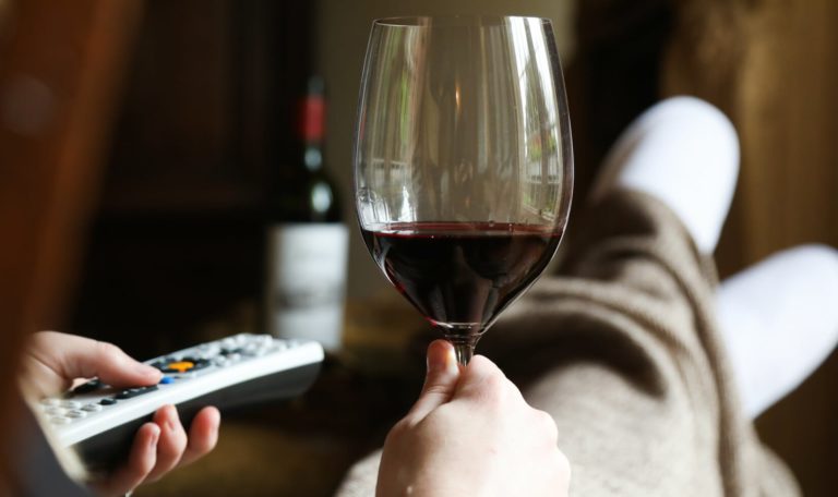 hands holding a glass of Jordan Winery Cabernet and a TV remote, with legs covered by a blanket in the background