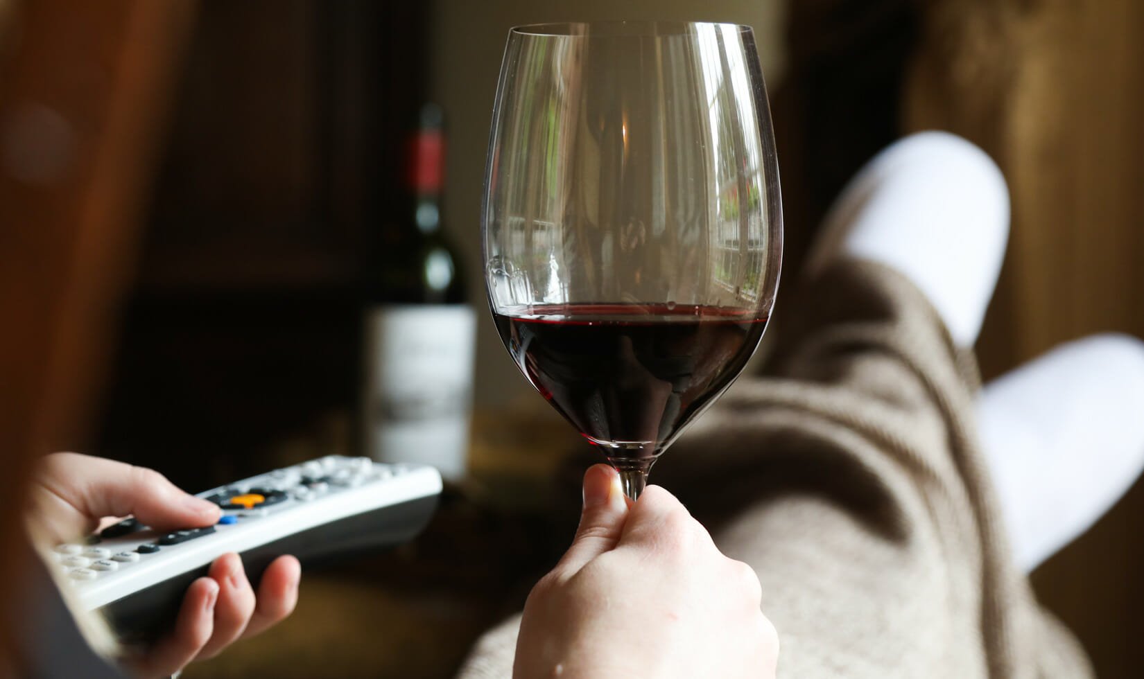 hands holding a glass of Jordan Winery Cabernet and a TV remote, with legs covered by a blanket in the background