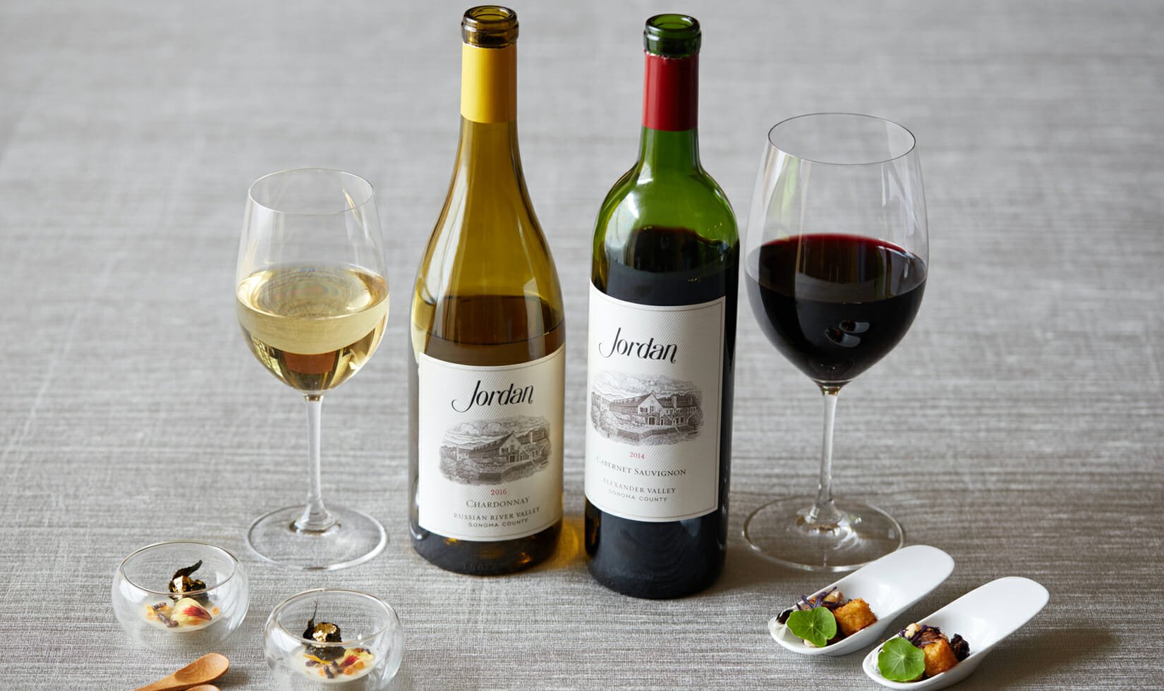 a bottle of Jordan Winery Chardonnay and Jordan Winery Cabernet next to two poured glasses of wine and small food bites