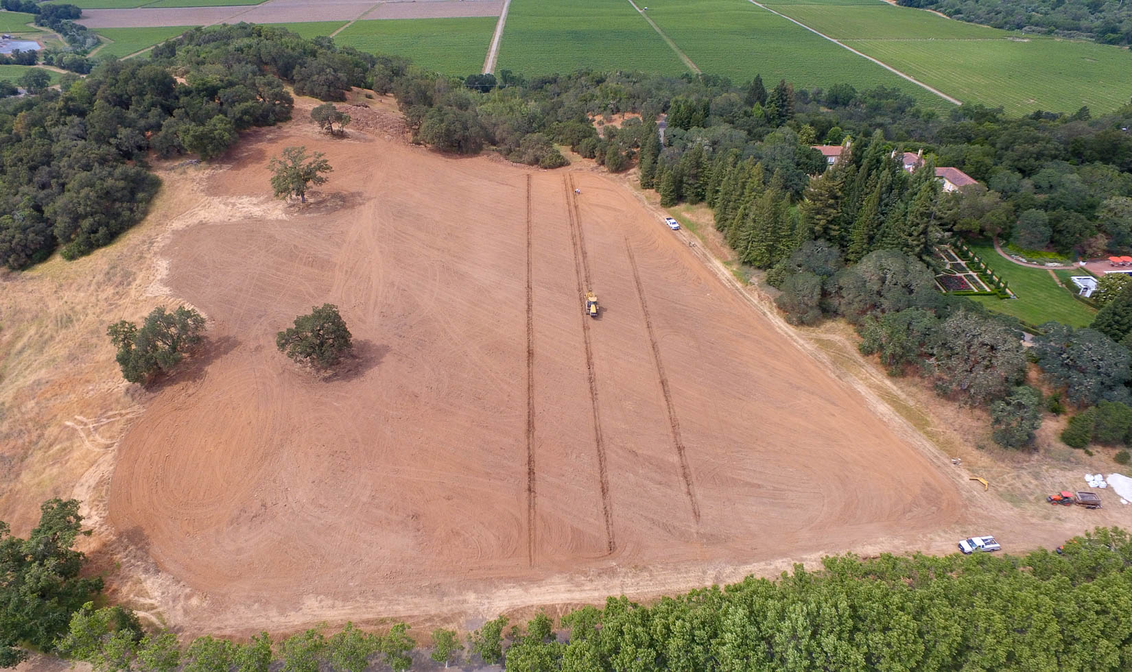 Aerial view of a tractor deep ripping vineyard soils for new Chateau Block at Jordan Winery. The dirt is shaped like a baseball field surrounded by trees.