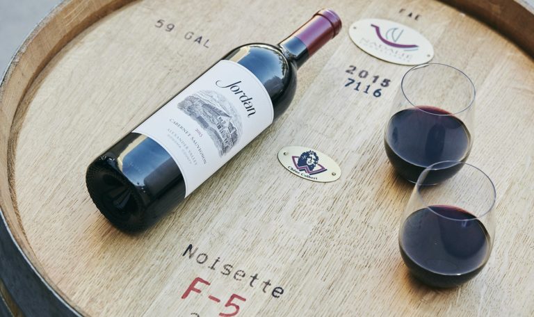 a bottle of 2015 Jordan Winery Cabernet beside two poured glasses of Cabernet on French oak barrel head