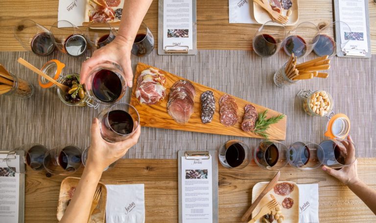 hands toasting with red wine in glasses over table setting for Jordan Winery Chateau Block Tasting with charcuterie and a Jordan Cabernet flight