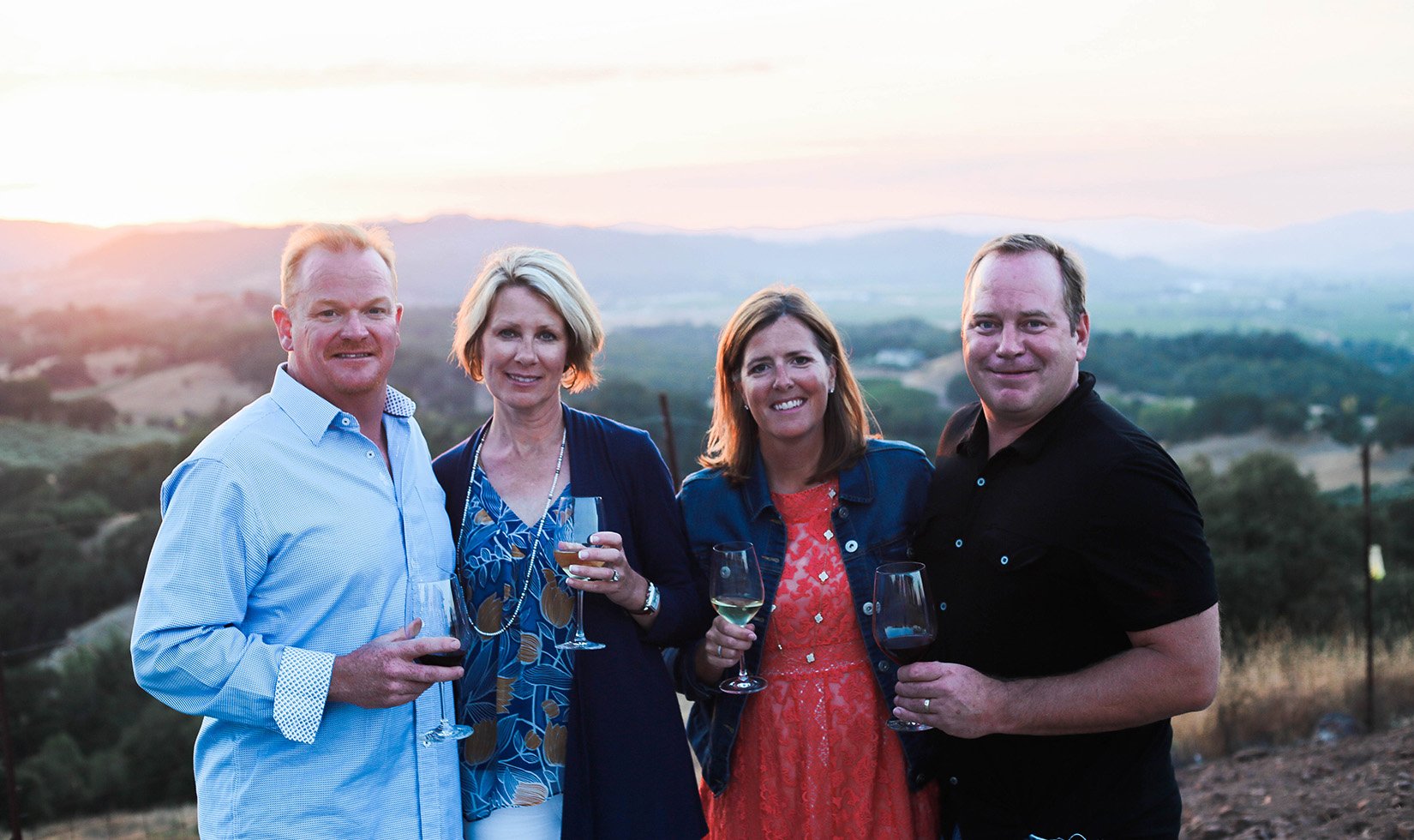 4 guests at Vista Point at Jordan Winery in wine country casual attire. The men are wearing button down shirts. The left woman is in a flowy tank top and cardigan while the woman on the right is in a lace floral dress with a denim jacket.