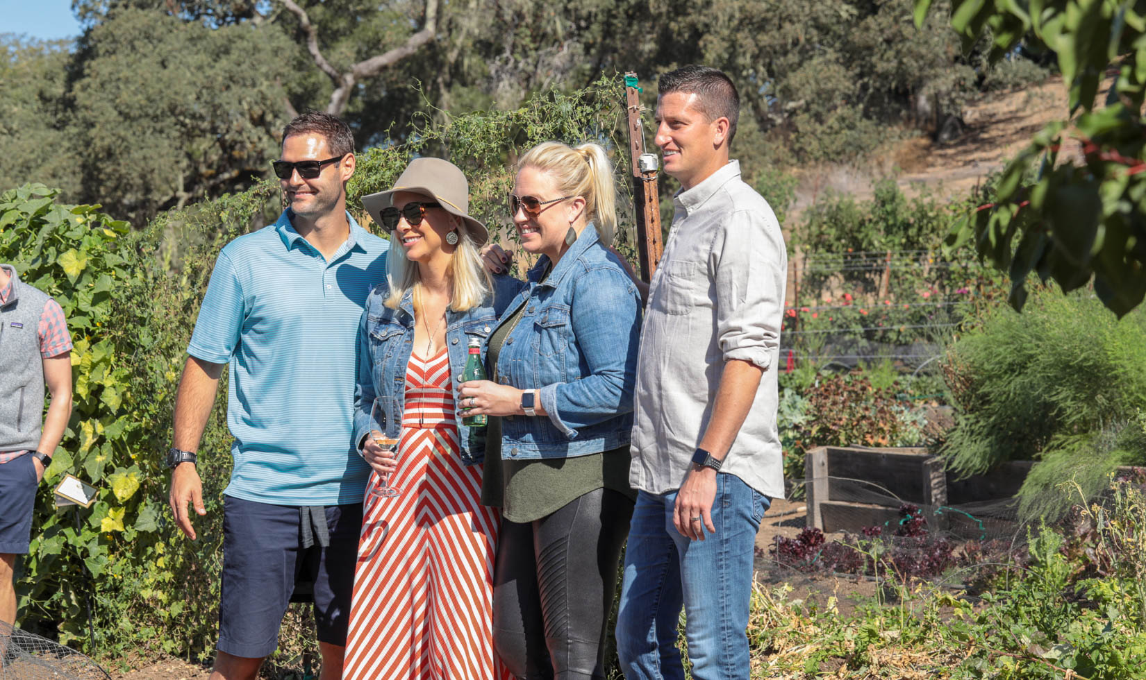 4 guests in the garden at Jordan Winery in wine country casual attire. The man on the left has a blue polo and shorts. Both women have denim jackets. The man on the right has a button down and jeans.