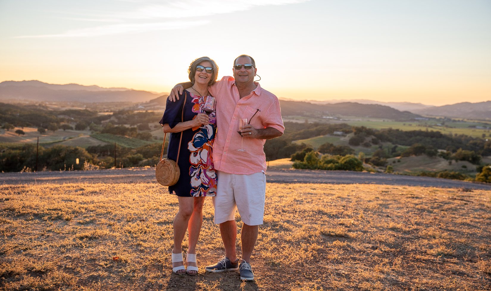 A couple at Vista Point at Jordan Winery in wine country casual attire. She's wearing an abstract floral dress and white sandal heels. He's in a salmon colored button down and shorts. They both have sunglasses and glasses of red wine. The sun sets behind them.