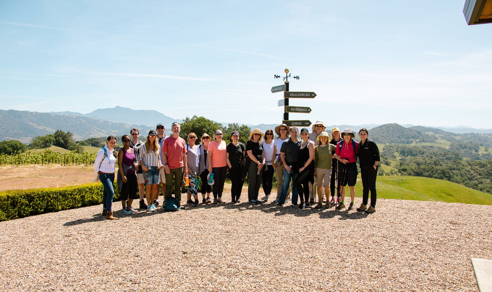 20 guests standing in front of a black and gold direction post. Most are in athletic outdoorsy clothes with sunglasses. Behind them are green vineyards and hills.