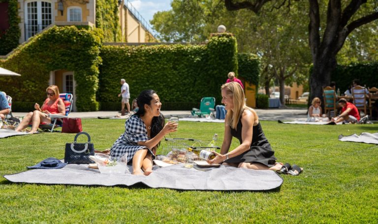 2 women laughing on a picnic blanket on Jordan Winery's lawn surrounded by other people on blankets. The woman on the left is wearing a black and white gingham plaid dress and holding a mason jar of chardonnay. The woman on the right is in a short black dress and pouring a jar of white wine.