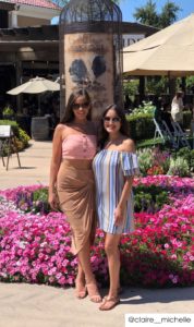 Two girls smiling in front of a winery wearing wine country casual clothing. The left one has a pink cropped tank top and beige knee length skirt. The right one has a vertical striped blue off-the-shoulder dress. Both wear sunglasses.