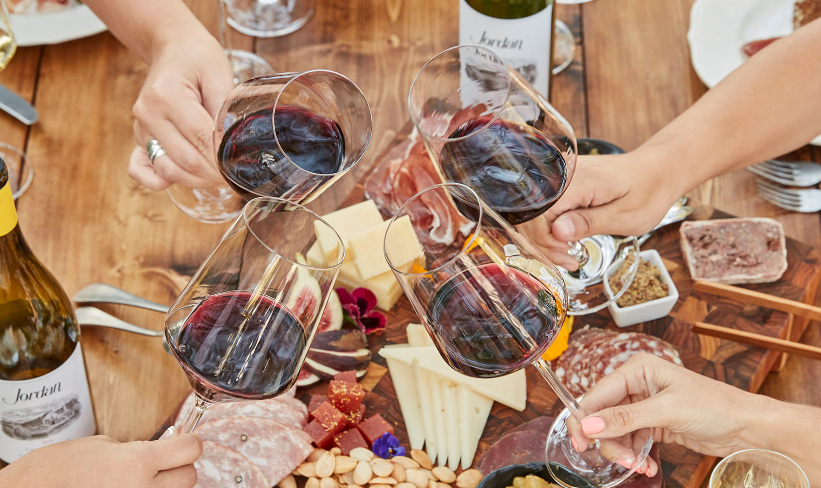 Hands toasting with glasses of Jordan Cabernet Sauvignon with food in the background