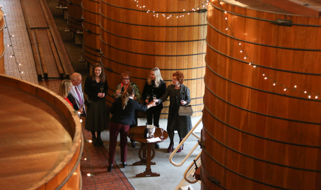 group of people standing in large oak tank room with christmas lights hung. They are toasting with wine glasses.