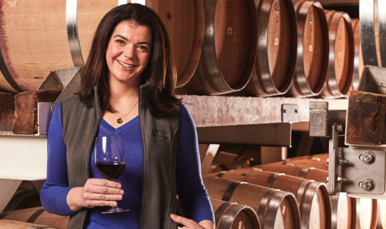 Winemaker Maggie Kruse with a glass of Jordan Cabernet Sauvignon, standing in front of barrels