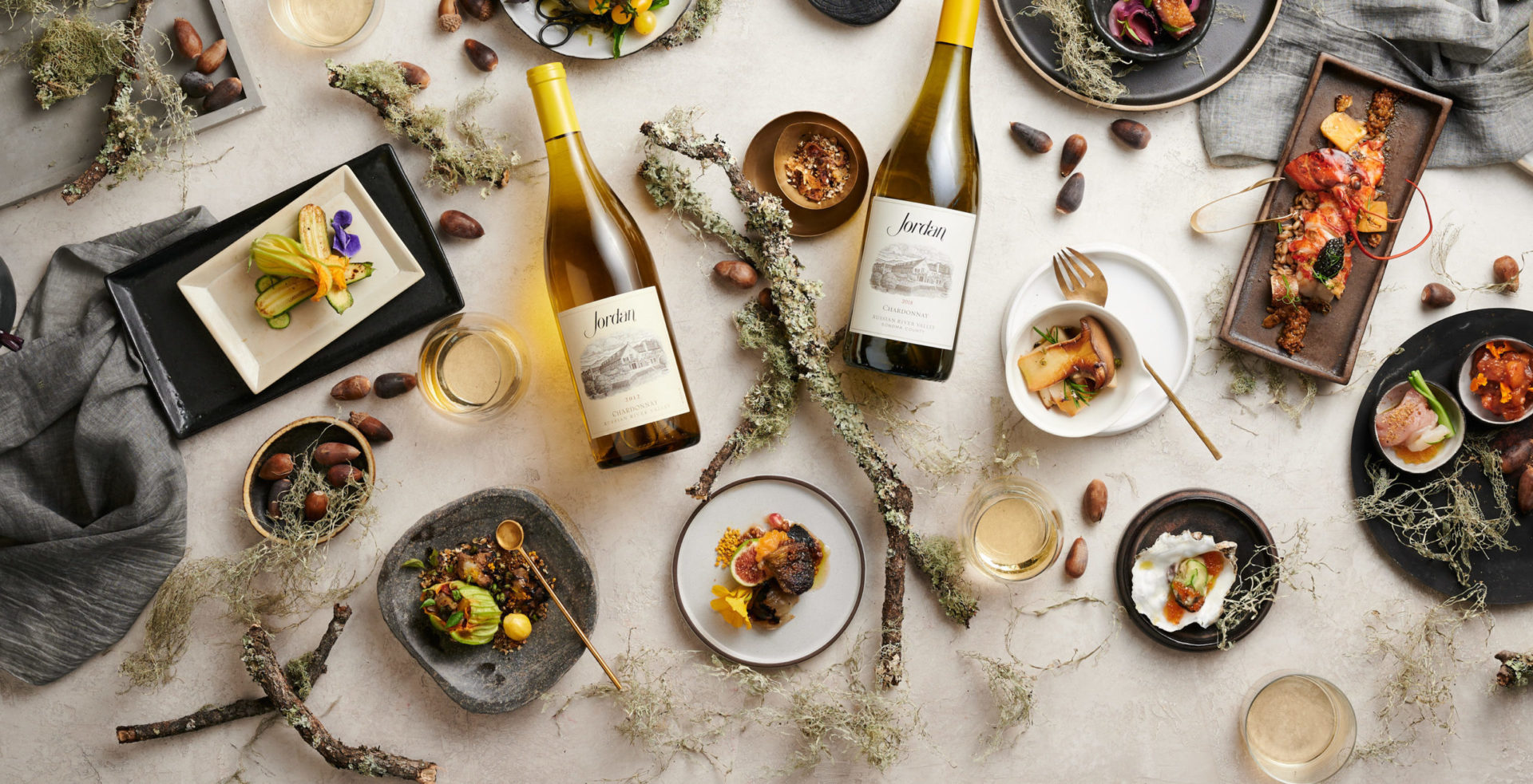Bottles of Jordan Chardonnay on a tabletop with food, linens, branches and acorns