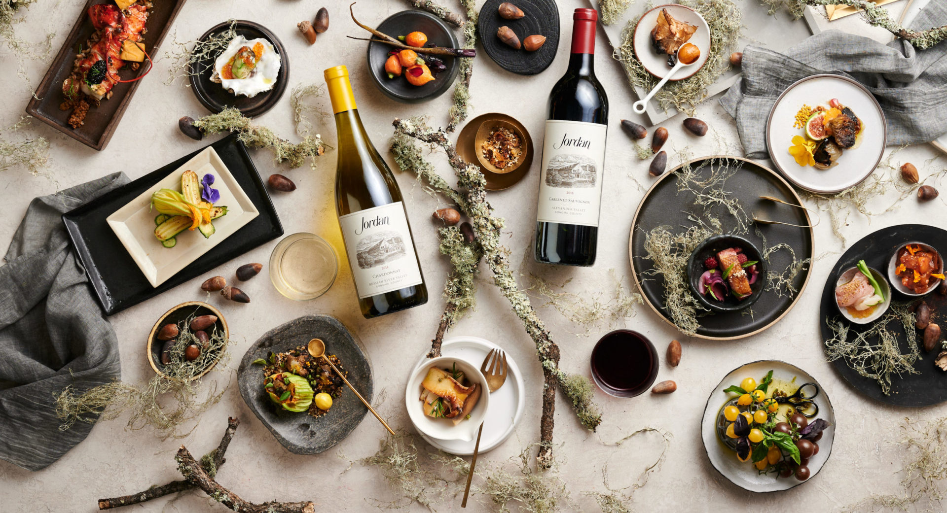 Bottles of Jordan Cabernet Sauvignon and Jordan Chardonnay on a tabletop with food, linens, branches and acorns