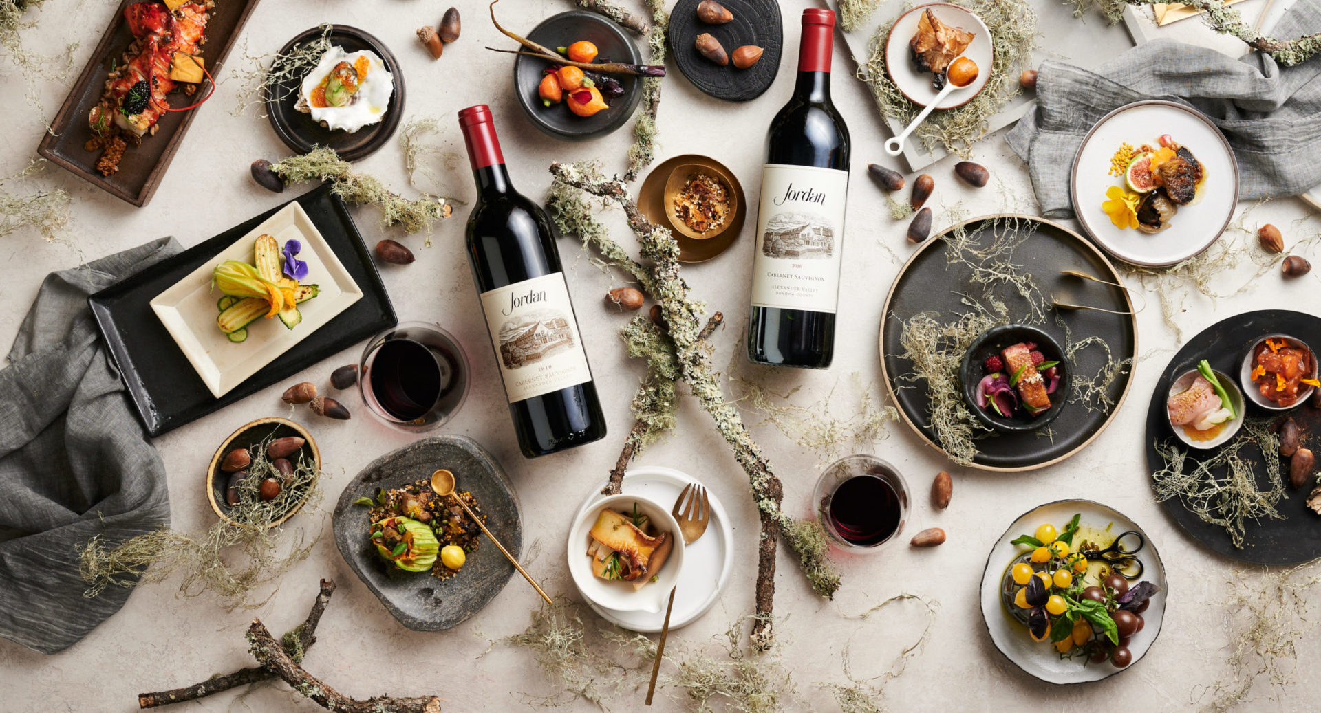 Bottles of Jordan Cabernet Sauvignon on a tabletop with food, linens, branches and acorns