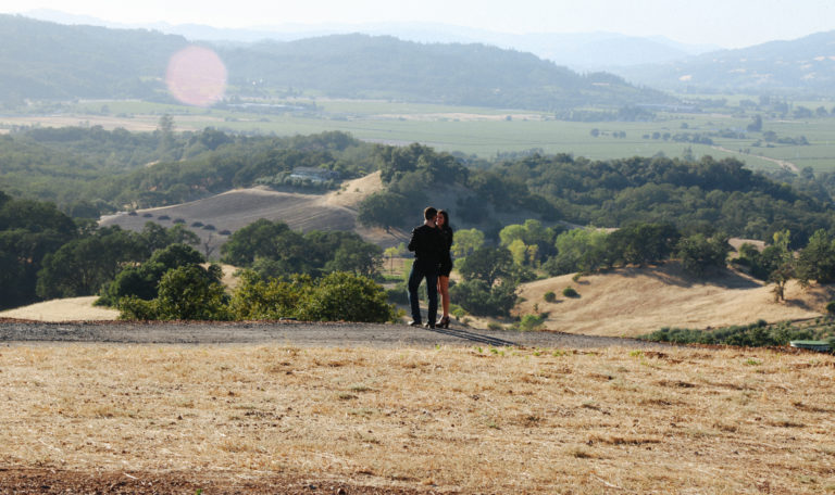 A man and woman embrace while overlooking golden rolling hills, oak trees, green vineyards, and mountains.