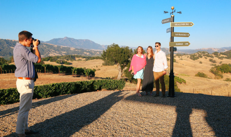 A man taking a picture of 2 women and a man next to a black and gold direction post. The 3 signs pointing right, from top to bottom, read "Healdsburg", "San Fransisco", and "Hawaii." Behind them are rolling golden hills and a vineyard on the left.