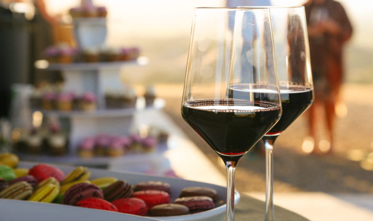 Close-up of 2 glasses of red wine to the right of a platter of yellow, red, and purple macarons. Blurred in the back left is a tiered cupcake stand.