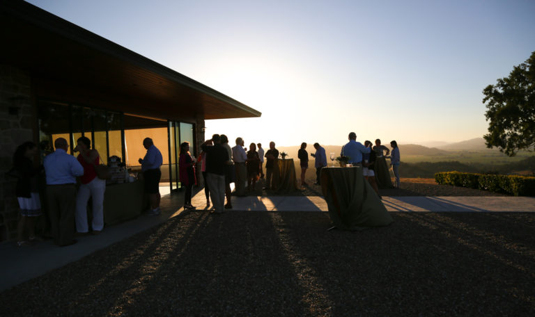 Guests silhouetted by the sunset overlooking vineyards. They stand outside a short modern building with floor to ceiling windows and sliding glass doors.