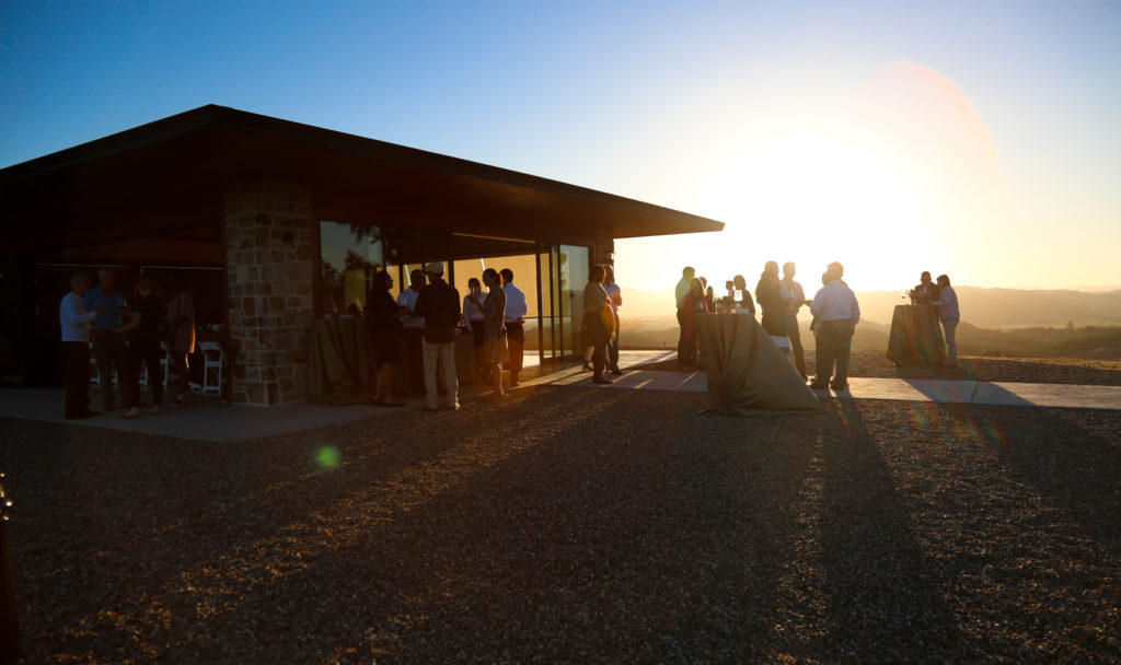 Guests silhouetted by the glowing sunset. They stand outside a short modern building with floor to ceiling windows, sliding glass doors, and cobblestone corners.