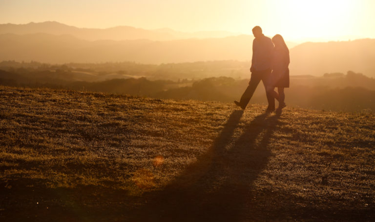 2 people silhouetted by the golden sunset walking left up a hill.