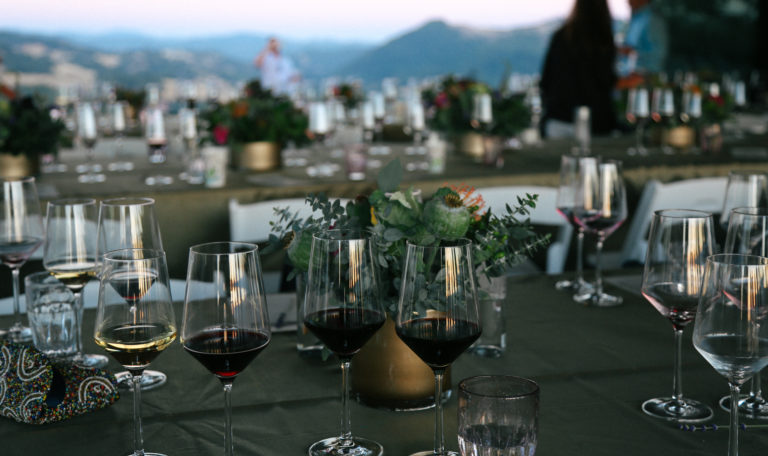 Partially empty glasses of red wine sit on a dark olive green table outside at twilight. An identical table and mountains are in the background.
