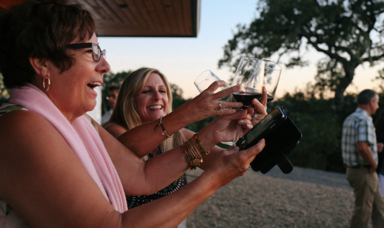 2 women laughing, clinking 3 glasses of red wine together, and taking a picture of the sunset they're viewing.