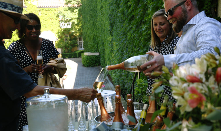 A bearded man in sunglasses pours a glass of champagne for an older man holding out his glass over the champagne station. 2 women look on smiling with their glasses. The courtyard walls are covered in dense ivy.
