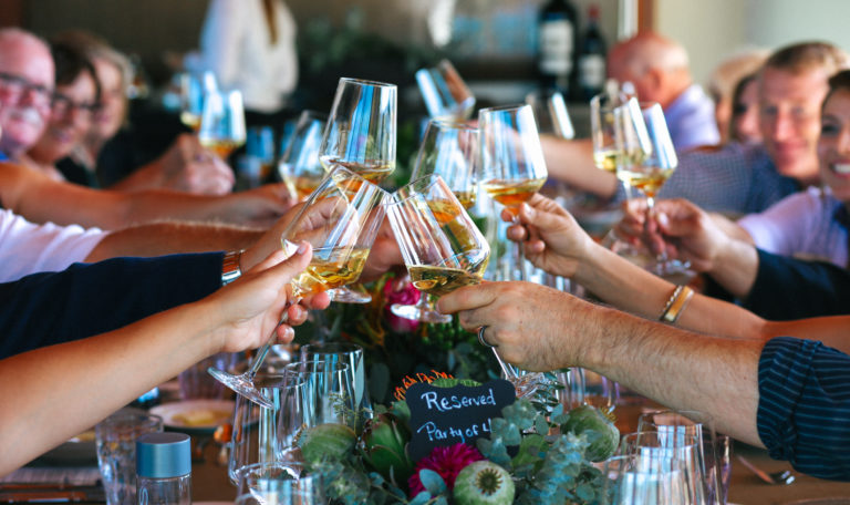 Guests' hands clinking together glasses of white wine. On the table in a bouquet, a sign reads "Reserved Party of 4."
