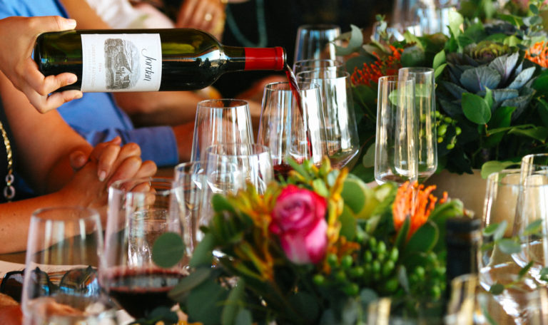 Multiple wine glasses between vibrant table bouquets. From the left a waiter's hand pours a bottle of Jordan's Cabernet Sauvignon.
