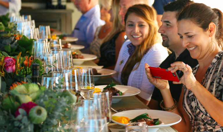 Guests just served a fancy steak entree. The woman on the right smiles and takes a picture of her dish with a red iPhone; another woman looks on smiling. Going down the table are multiple wine glasses and vibrant green bouquets.