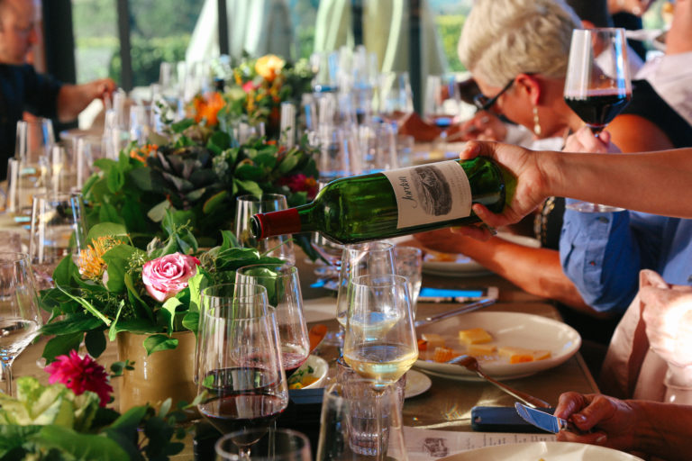 A waiter's hand coming from the right to pour a bottle of red wine. On the table are multiple wine glasses and small green bouquets.