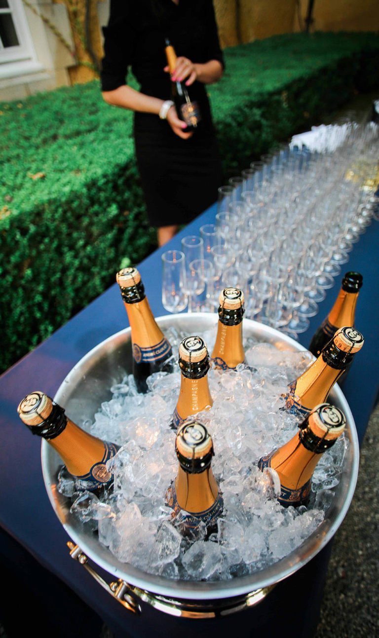 Overhead shot of a champagne ice bucket with 7 bottles. On the table behind are rows of champagne glasses and a server.