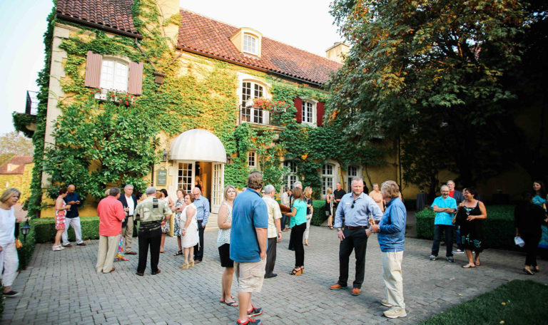 Guests socializing in the Jordan Winery courtyard. The building is yellow with white windows and a brick roof. It's exterior walls are completely covered in ivy and shrubbery. A large tree hangs in from the right.