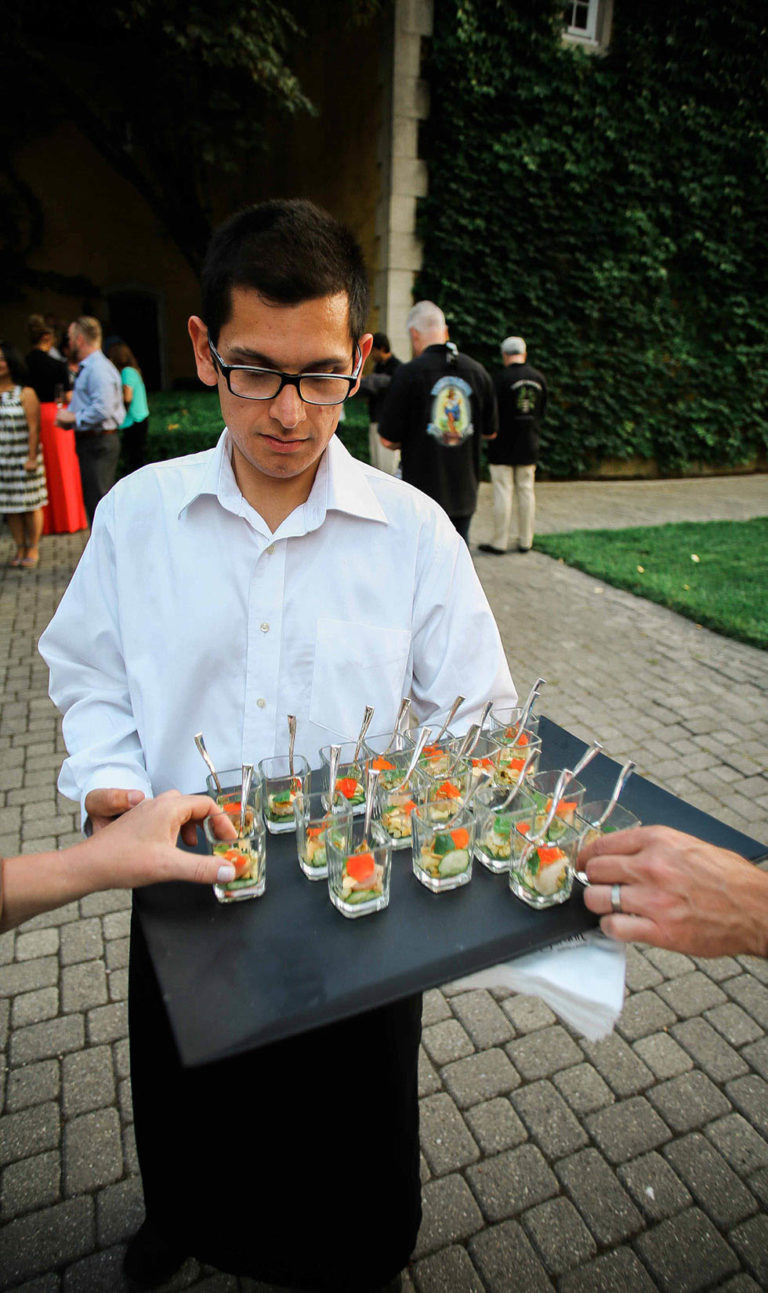 A server holding a black tray of appetizers in small glasses as 2 hands from either side reach to grab one.