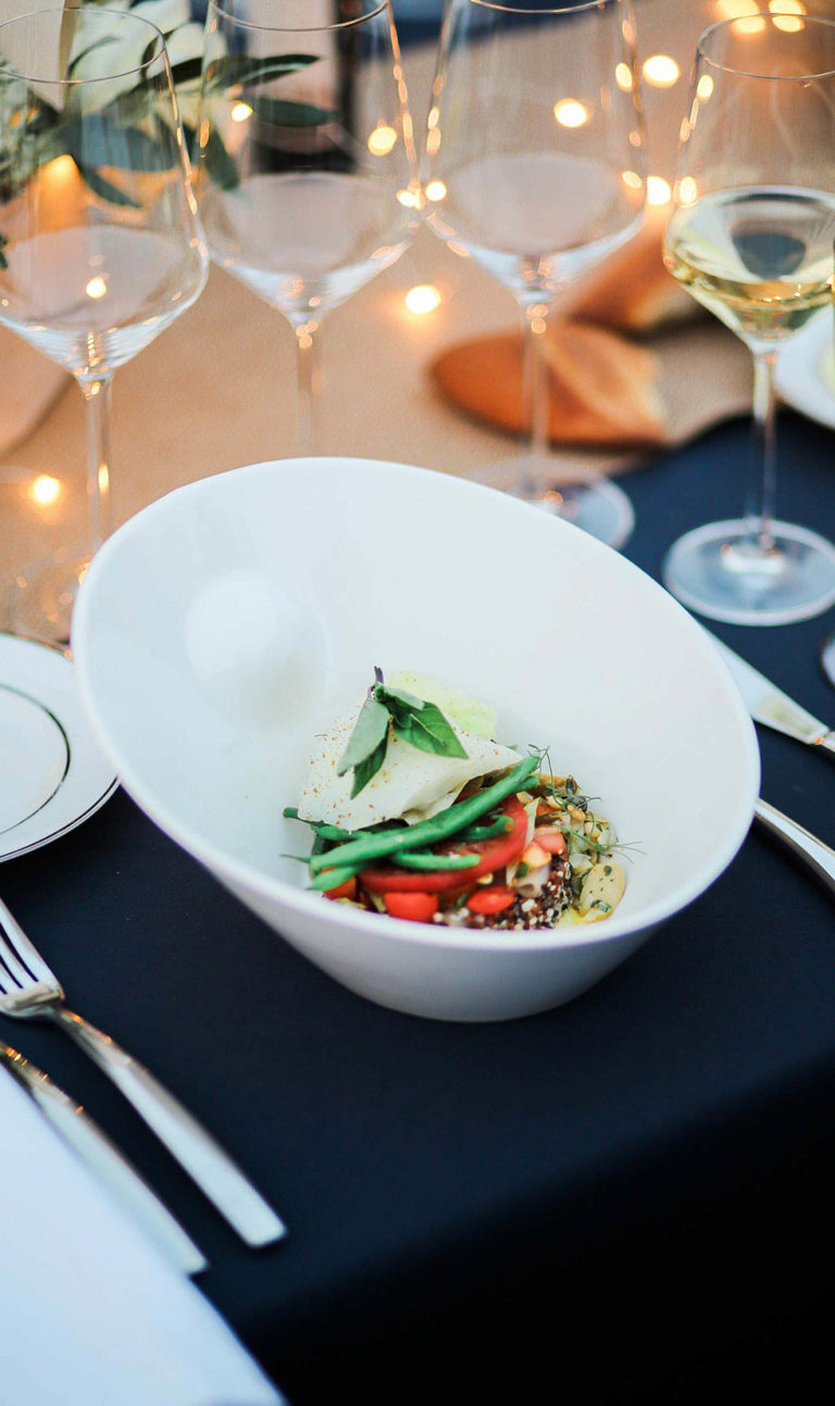 Mixed quinoa, tomato, and green bean vegetable entree in fine bowl surrounded by wine glasses.