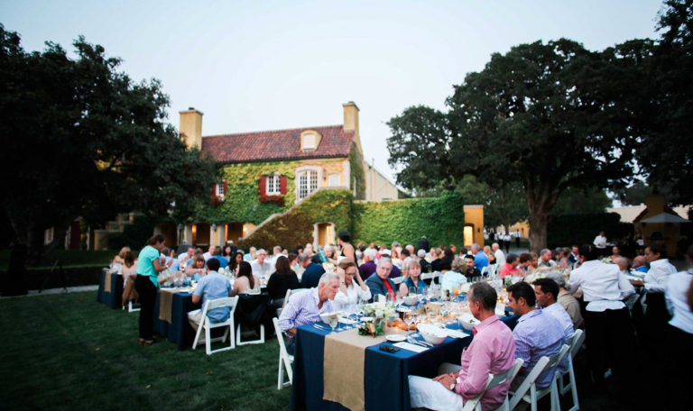 Guests seated at 3 long tables on Jordan Winery's lawn. There are formal place settings with navy blue table cloths and burlap runners. In the background, the sunset shines on Jordan's tasting room. The building is covered in ivy, has a brick roof, and white windows with red shutters.