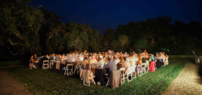 Guests seated at 3 long tables on Jordan Winery's lawn. There are formal place settings with navy blue table cloths and burlap runners. There are oak trees in the middle ground with a starry blue night sky above.