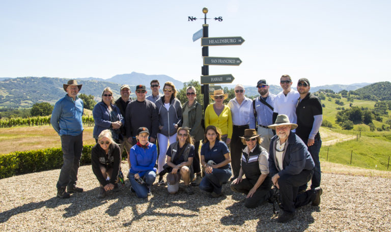 A group of 18 people in outdoorsy athletic attire smile in front of a black and gold direction post. The signs pointing right, "South" according to the arrows, read "Healdsburg", "San Fransisco", and "Hawaii." Behind them are green vineyards, mountains, and a trail.