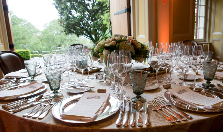 Formal dining settings at a round table with multiple wine and champagne glasses for tastings. The table is in front of large, open, double wooden doors and a bouquet of hydrangeas and roses sits at the center.