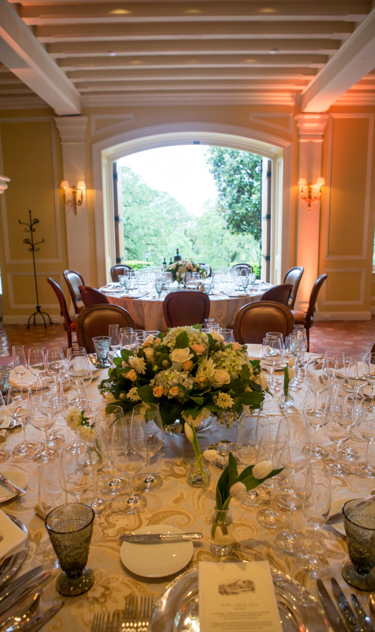 Formal dining settings at a round table with multiple wine and champagne glasses for tastings. The table is inline with an identical dinner table in front of large, open, double wooden doors and a bouquet of hydrangeas and roses sits at the center of each.